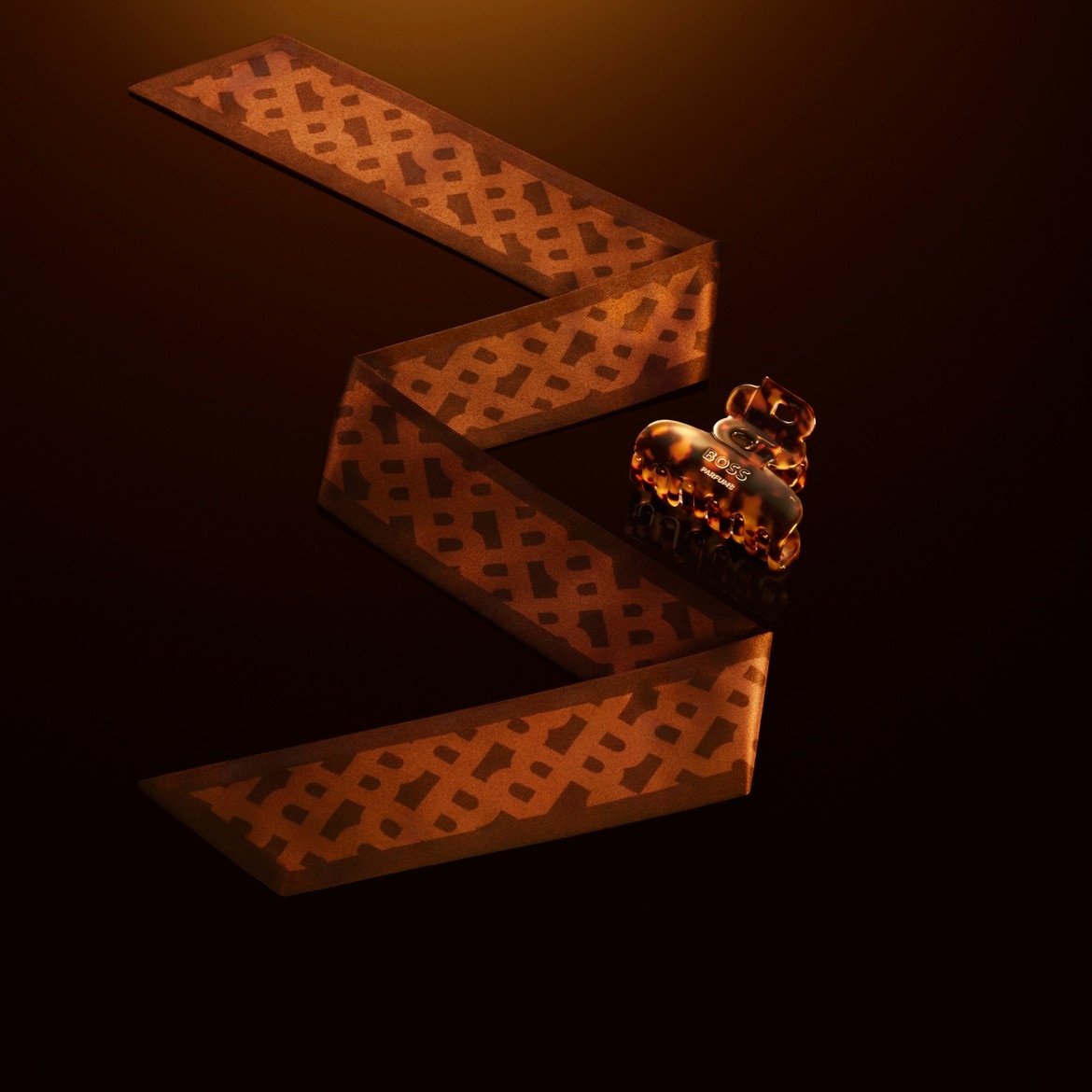 Auf einem dunklen Untergrund liegt ein gewundenes Band mit einem braunen Muster, daneben eine Haarspange mit dem Schriftzug BOSS Parfums.