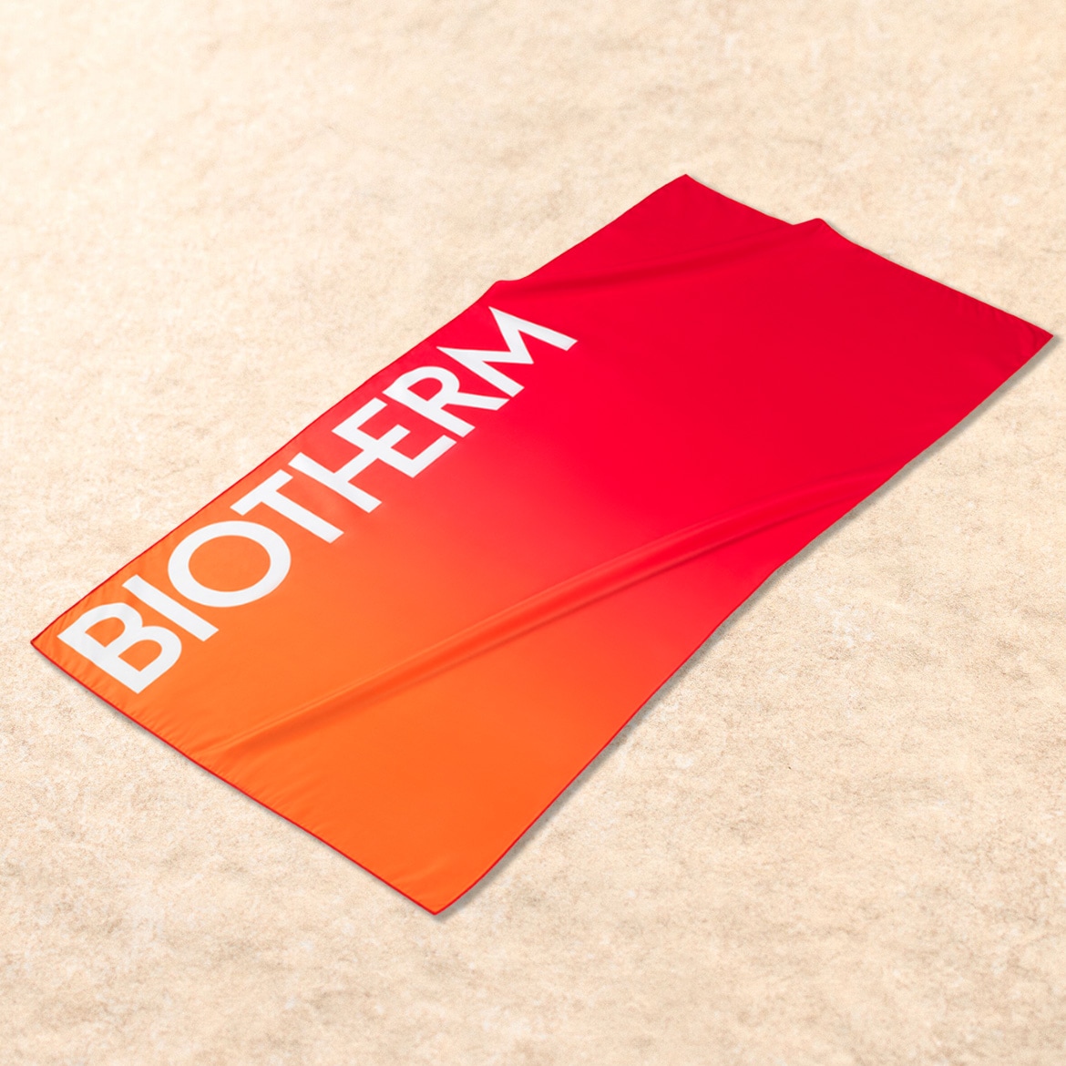 Ein leuchtend rot-orangefarbenes Strandtuch mit dem weißen BIOTHERM-Logo liegt auf hellem Sand und lädt zum Entspannen ein.