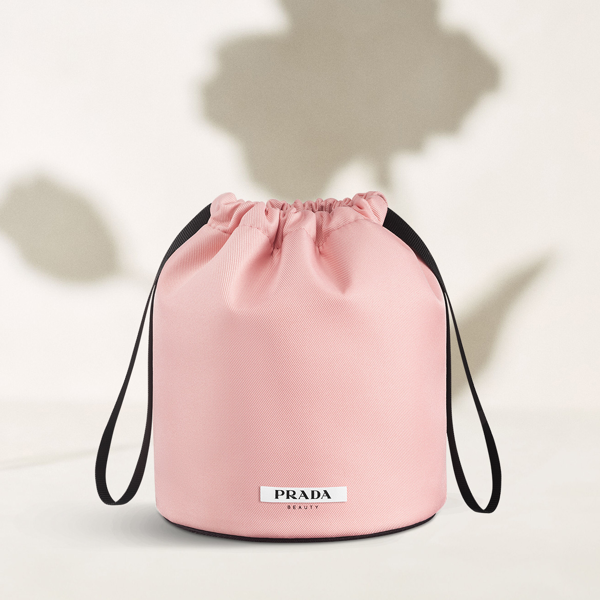 Ein rosafarbener Prada Beauty Beutel mit Kordelzug und schwarzem Band, der auf einer hellen Oberfläche steht und von Schattenblumen umgeben ist.