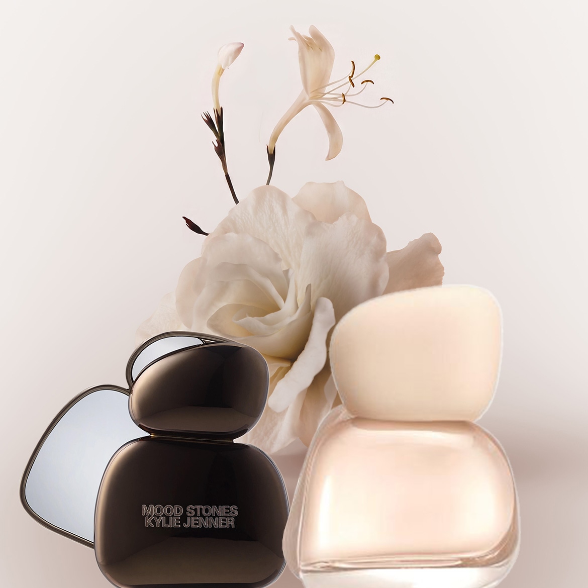 Zwei Parfümflaschen, eine schwarze mit der Aufschrift "MOOD STONES KYLIE JENNER" und eine hellbeige, stehen vor einem Hintergrund aus weißen Blüten.
