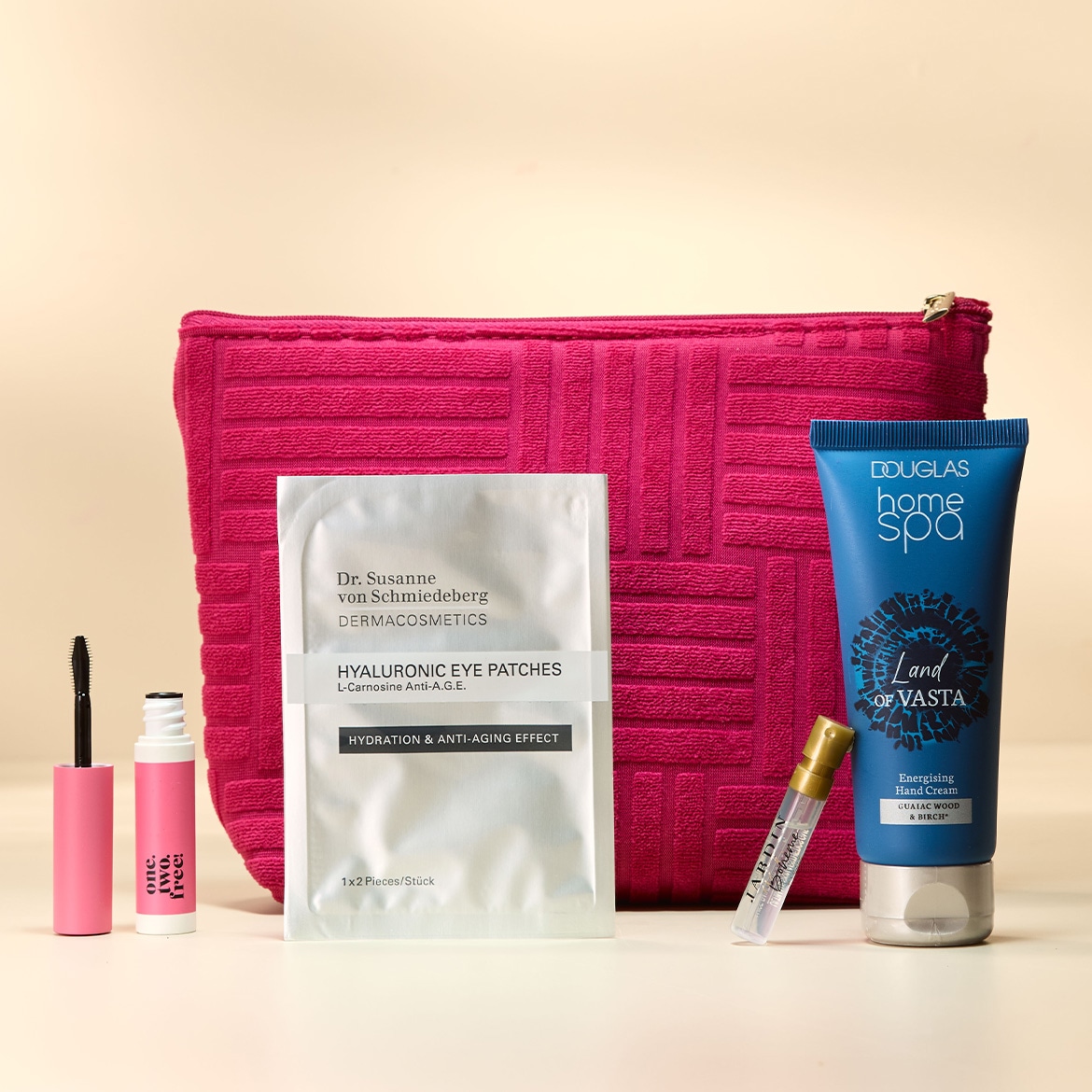 Das Bild zeigt eine Kosmetiktasche mit einem Reißverschluss, daneben eine schwarze Mascara und eine pinkfarbene Tube mit der Aufschrift "one. two. free!".