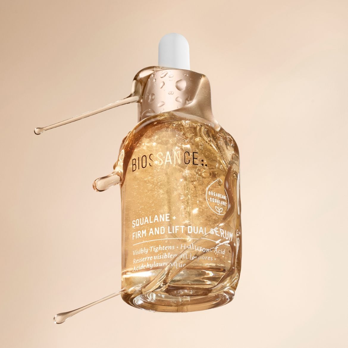 Das Biossance Squalan + Firm and Lift Dual Serum in einer durchsichtigen Glasflasche mit Pipette, umgeben von goldenen Flüssigkeitstropfen, verspricht eine straffere und sichtbar verjüngte Haut.