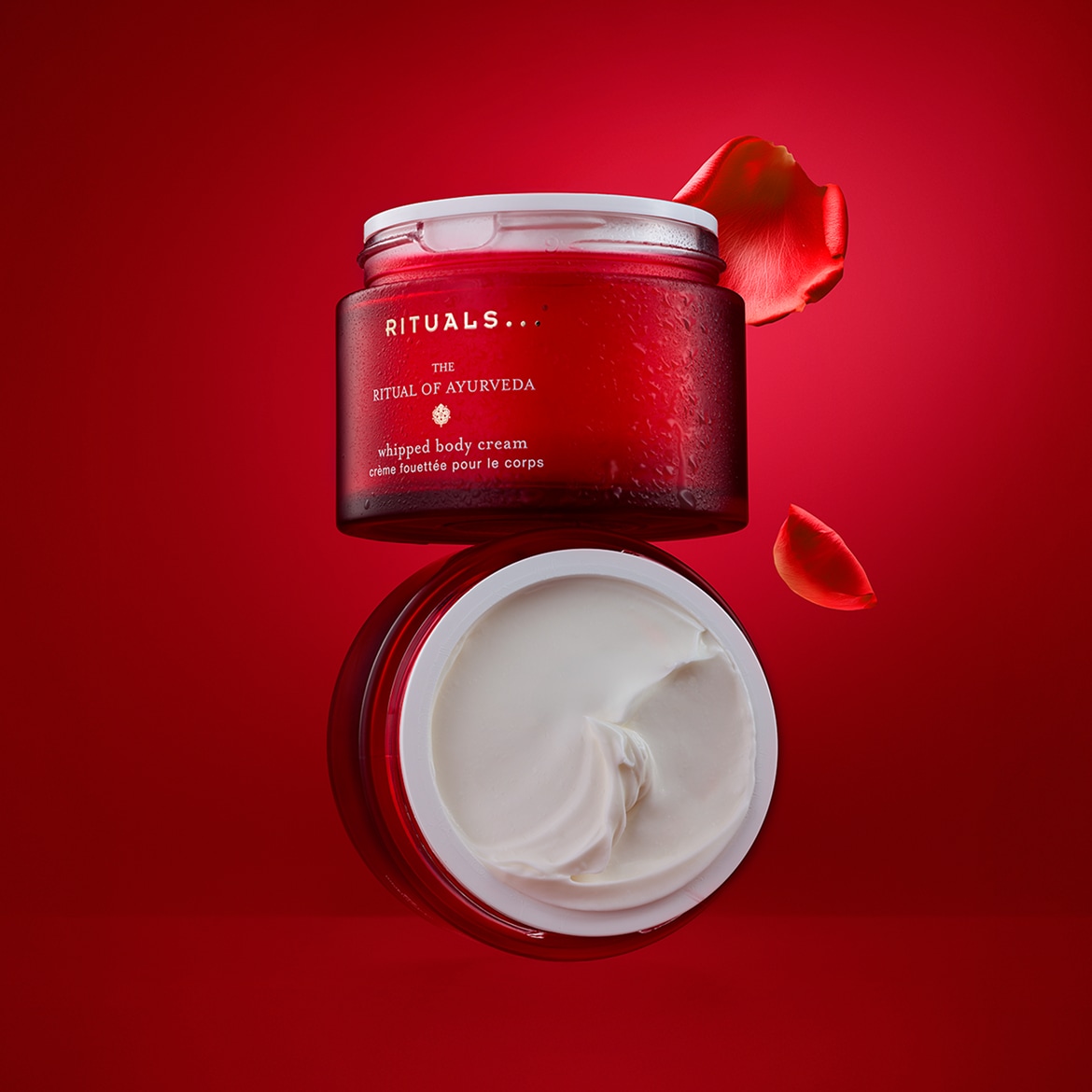 Zwei rote Tiegel Rituals The Ritual of Ayurveda Whipped Body Cream stehen übereinander, daneben liegen rote Blütenblätter.