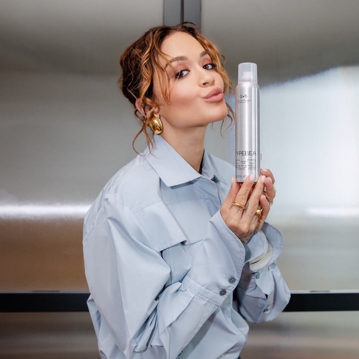 Eine Frau mit gewelltem, braunem Haar und goldenen Ohrringen hält eine silberne Dose Trockenshampoo der Marke Typedbea G+5 in ihren Händen.