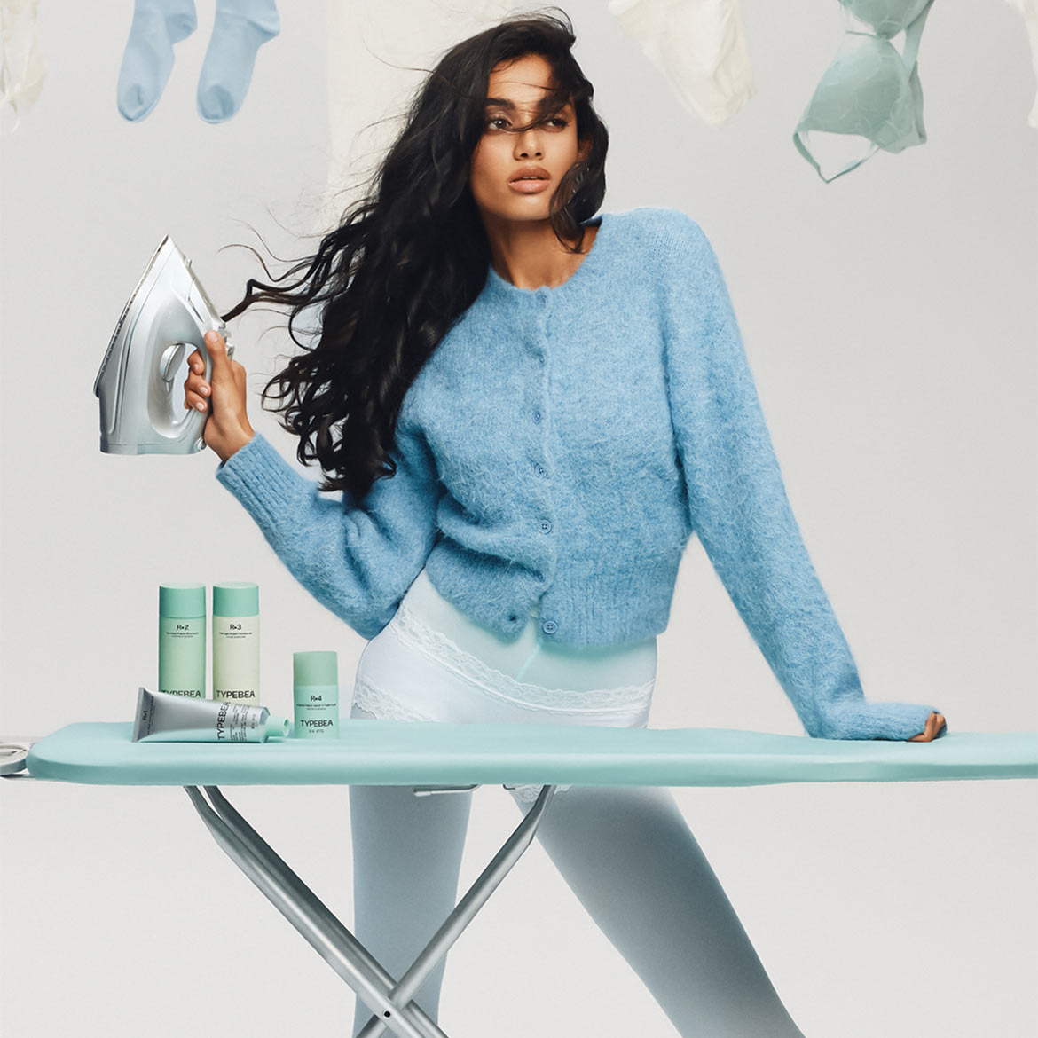 Eine Frau mit wallendem, dunklem Haar hält ein Bügeleisen in der Hand, während sie an einem Bügelbrett mit Hautpflegeprodukten der Marke TYPEBEA steht.