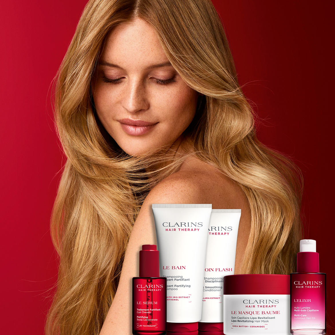 Eine Frau mit langen, blonden Haaren blickt mit geschlossenen Augen nach unten, umgeben von einer Auswahl an Clarins Hair Therapy Produkten, darunter ein Shampoo, Conditioner, Serum, Maske und Haaröl, alle in eleganten roten und weißen Verpackungen.