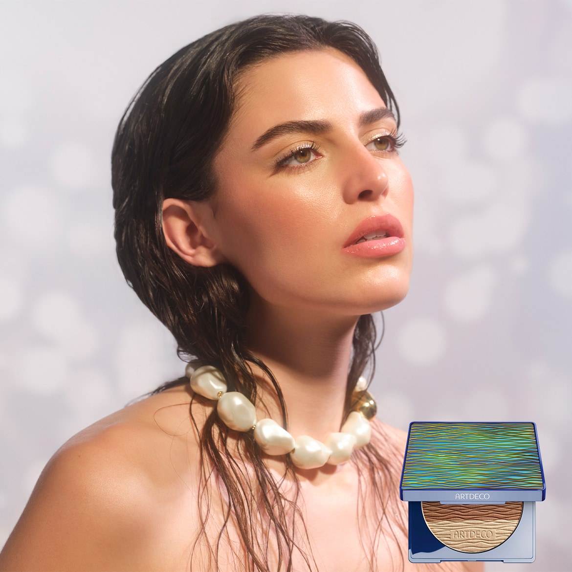 Eine Frau mit nassen, dunklen Haaren und einer Perlenkette blickt nach oben, neben ihr ein Artdeco-Puder mit blau-grüner Verpackung.