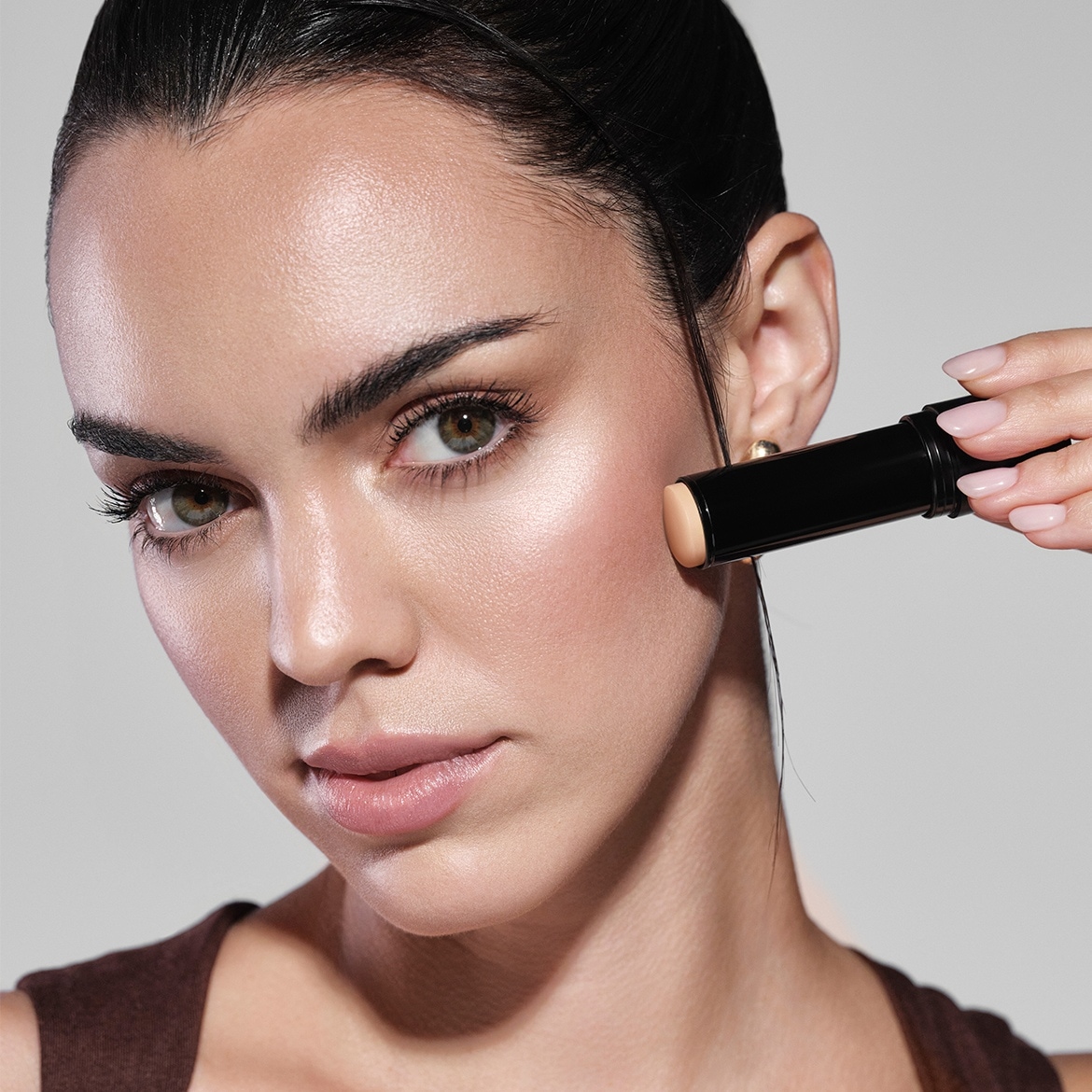 Eine Frau mit olivfarbenen Augen und zurückgekämmtem Haar trägt einen beigen Make-up-Stick auf ihre Wange auf.