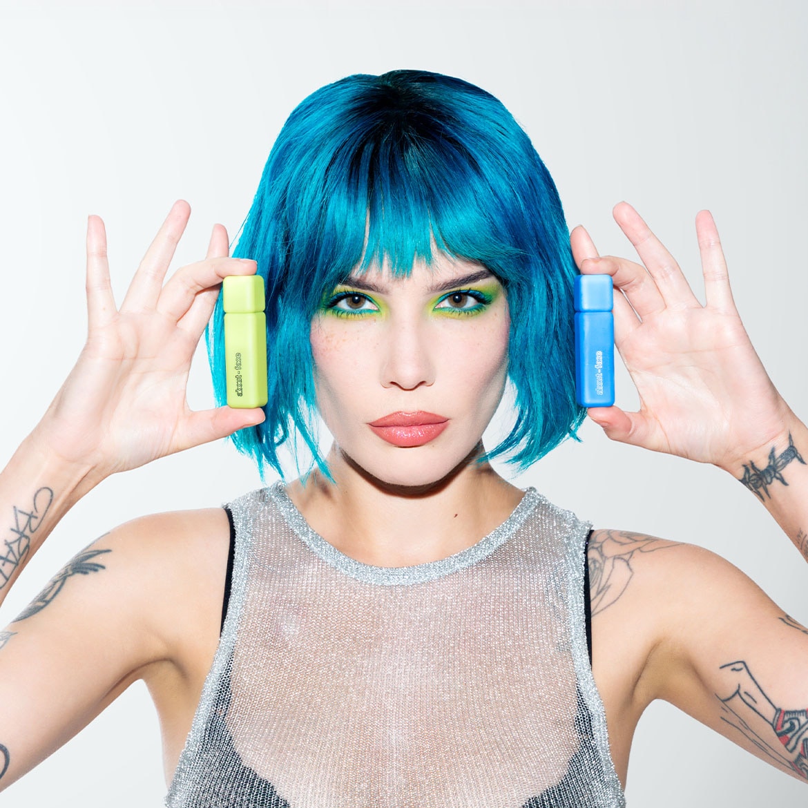 Eine Frau mit blauen Haaren und glitzerndem Oberteil hält in jeder Hand eine farbige Flasche, eine hellgrün und die andere blau.