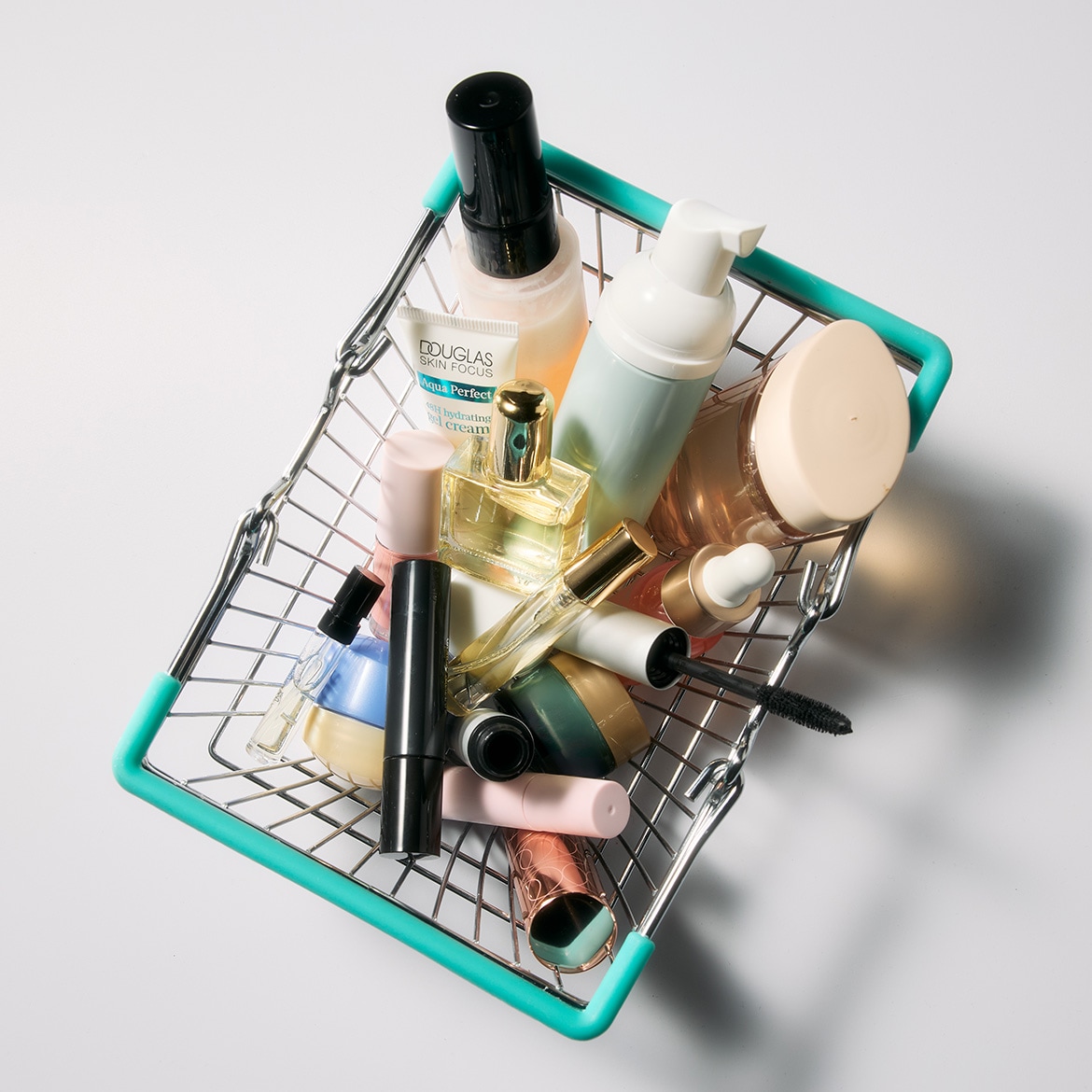 Ein silberner Einkaufskorb mit türkisfarbenen Griffen ist voll mit verschiedenen Schönheits- und Hautpflegeprodukten, darunter Flakons, Cremes und Make-up.