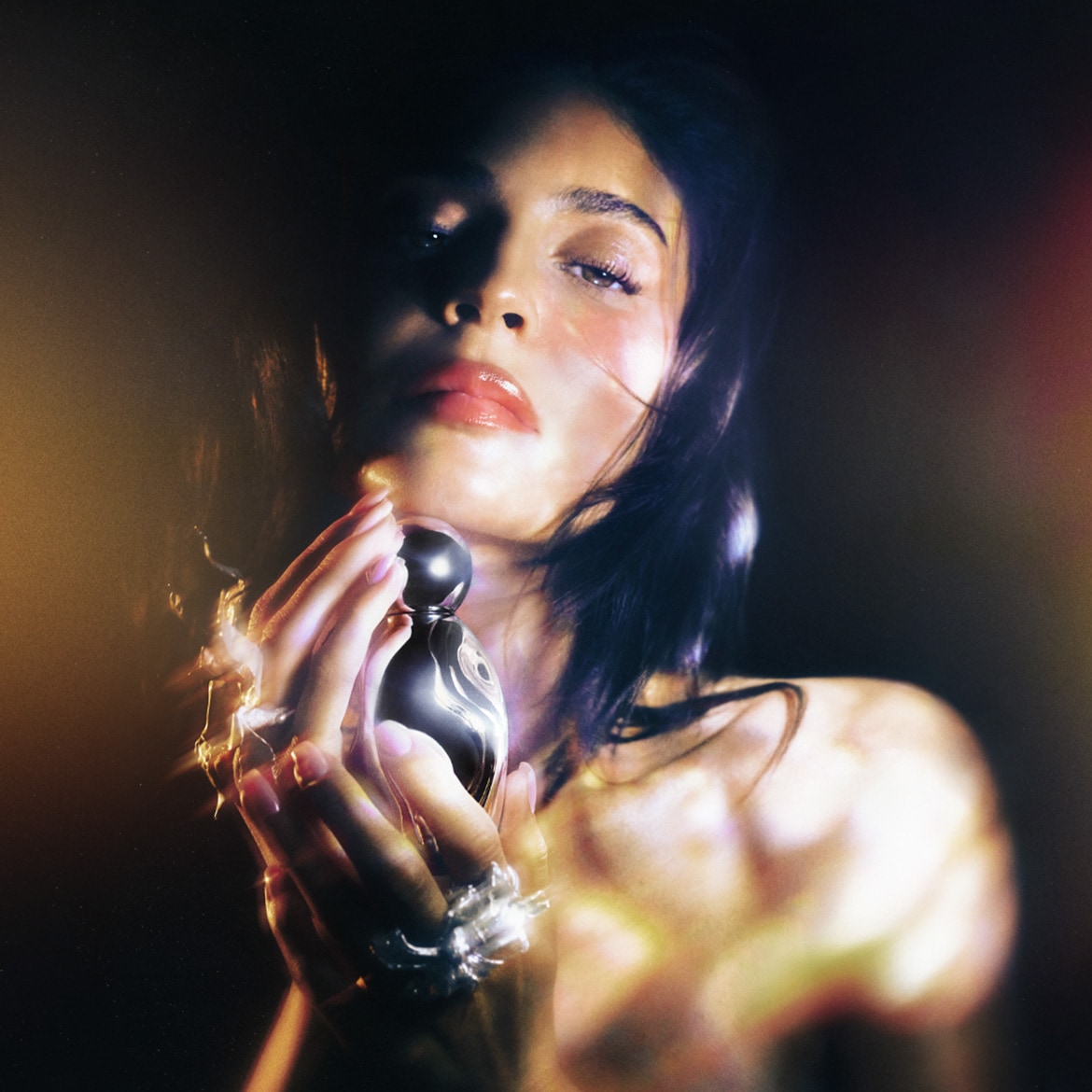 Eine Frau mit glänzenden Lippen hält einen schwarzen Parfümflakon mit silbernen Wirbeln in den Händen, die mit auffälligen Ringen geschmückt sind.