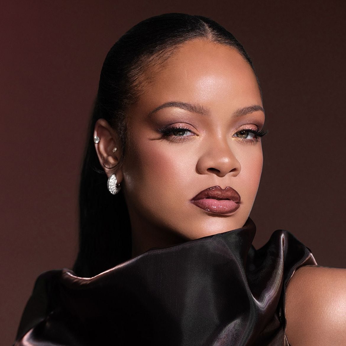 Rihanna trägt ein glänzendes Make-up mit braunem Lidschatten, glänzenden Lippen und einem schwarzen Oberteil, das ihre Schultern bedeckt.