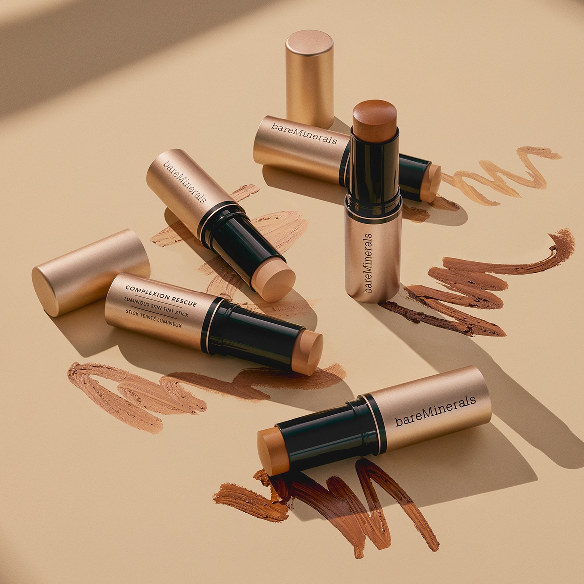 bareMinerals Complexion Rescue Luminous Skin Tint Sticks in verschiedenen Hauttönen, von hell bis dunkel, sind auf einem beigefarbenen Hintergrund angeordnet, mit Farbmustern, die die Vielfalt der Farbtöne zeigen.