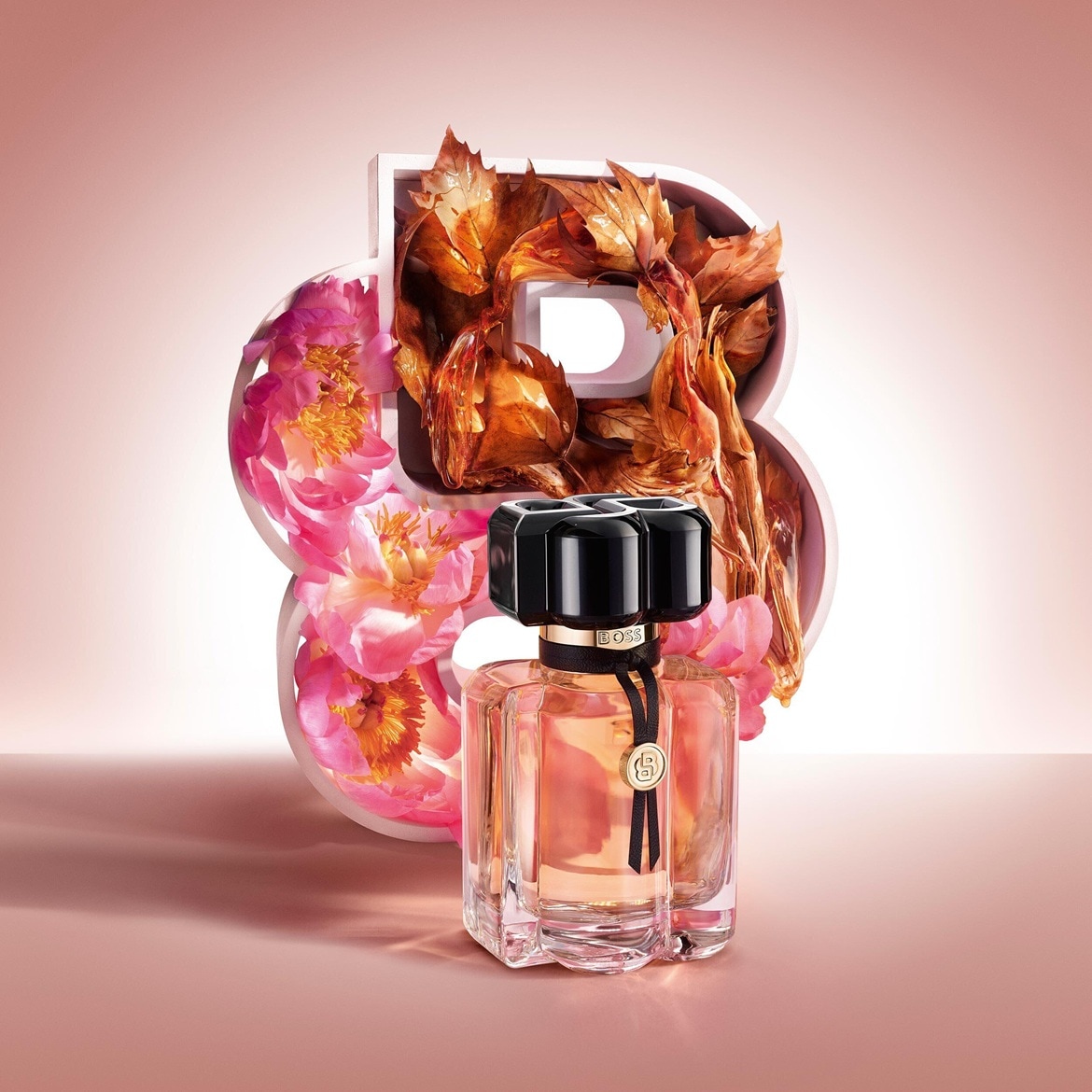 Ein rechteckiger, transparenter Flakon von Boss The Scent Elixir mit goldener Flüssigkeit, schwarzem Verschluss und schwarzem Band mit goldenem Logo steht vor einer großen weißen "B" mit rosa Blüten und braunen Blättern.