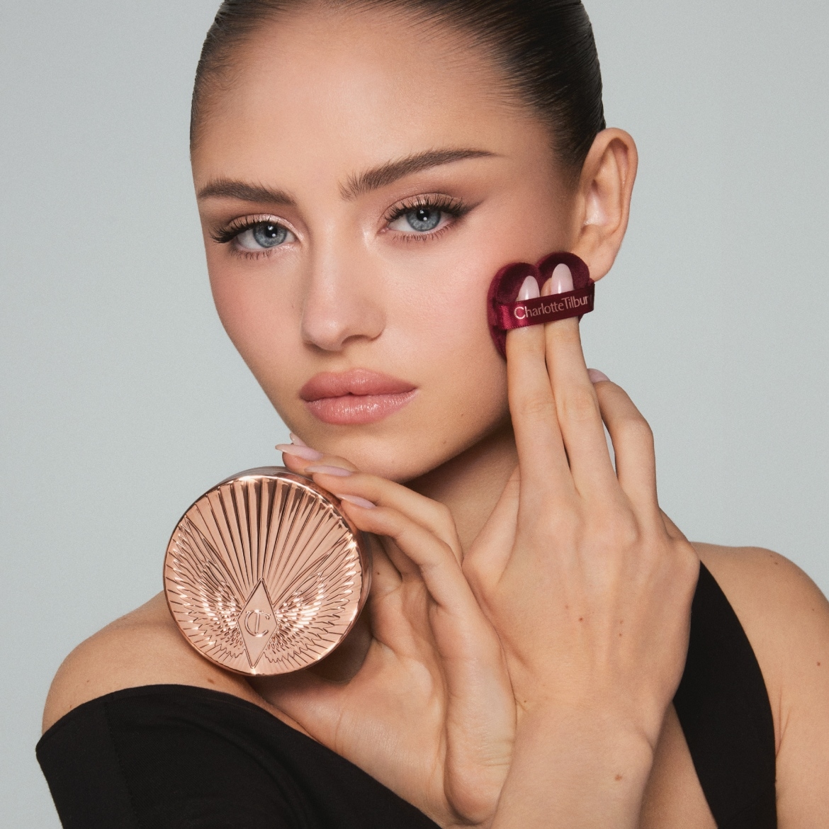 Eine Fraueinem schwarzen Oberteil hält eine runde, roségoldene Puderdose mit Flügelmuster in der linken Hand und tupft mit einem herzförmigen, weinroten Puderquaste mit der Aufschrift „Charlotte Tilbury“ auf ihre rechte Wange.