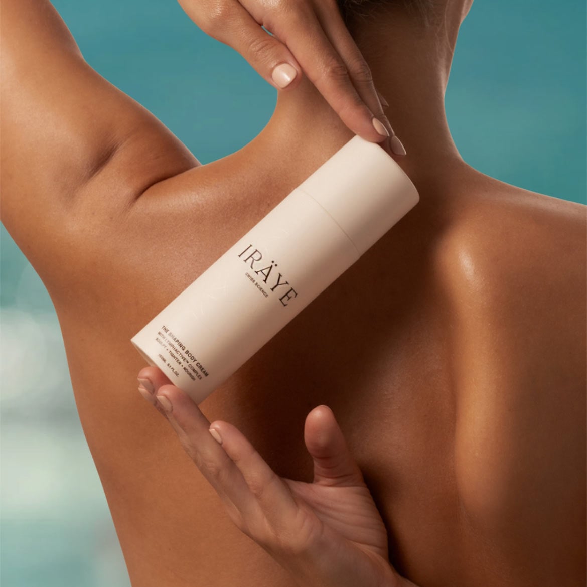 Eine Person hält eine weiße Flasche IRÄYE The Shaping Body Cream mit Lymphactive Complex, die die Haut formt, strafft und nährt, vor ihrem Rücken.