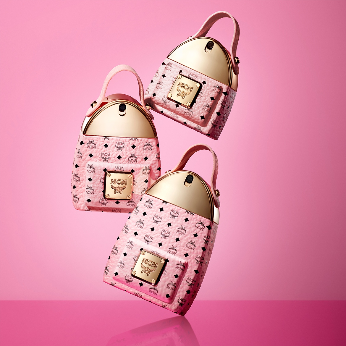 Drei MCM-Handtaschen in Rosa mit goldenen Akzenten schweben vor einem passenden rosa Hintergrund und zeigen das ikonische MCM-Logo.