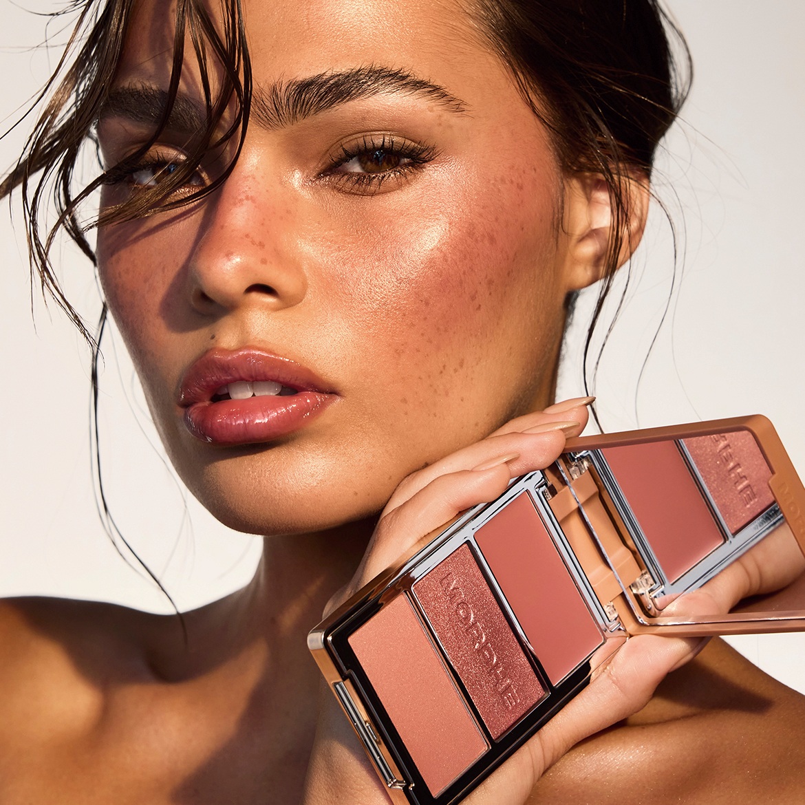 Nahaufnahme eines Models mit taufrischem Make-up, das eine Morphe-Palette mit Rouge- und Highlighter-Tönen in Pfirsich- und Rosatönen hält, um einen strahlenden Teint zu erzielen.