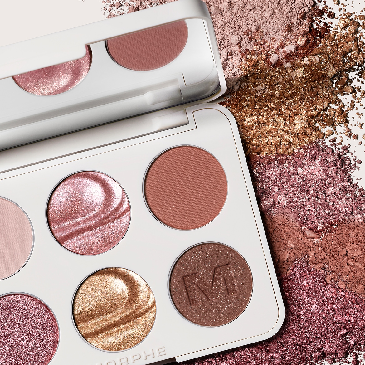 Nahaufnahme einer Morphe-Palette mit sechs Farbtönen in Rosa-, Gold- und Brauntönen, ergänzt durch loses, schimmerndes Puder in ähnlichen Farbtönen.