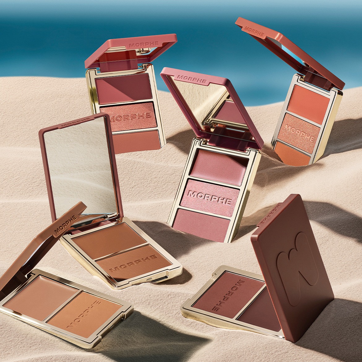 Eine Nahaufnahme von Morphe Rouge- und Bronzer-Paletten in verschiedenen Farbtönen, die auf sandfarbenem Untergrund mit blauem Hintergrund angeordnet sind und eine Auswahl an warmen Farbtönen für Wangen und Gesicht zeigen.