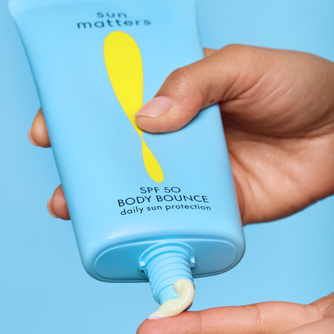 Eine Hand drückt eine hellblaue Tube mit der Aufschrift „sun matters SPF 50 BODY BOUNCE daily sun protection“ und einem gelben, tropfenförmigen Logo aus, wobei eine cremefarbene Sonnencreme auf einen Finger aufgetragen wird.