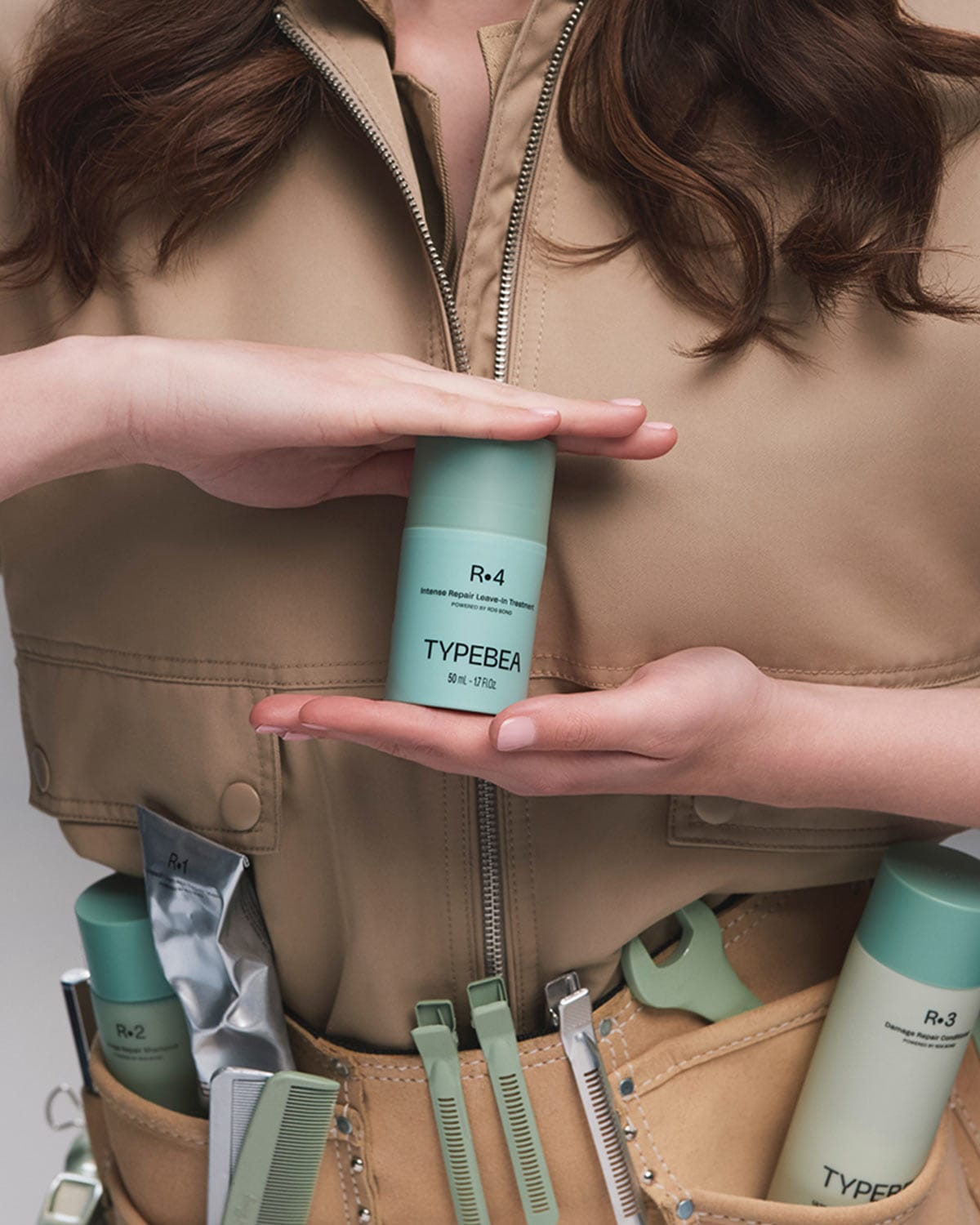 Eine Person in beigefarbener Kleidung hält eine mintgrüne Flasche TYPEBEA R.4 Intense Repair Leave-In Treatment in den Händen, während weitere Produkte und Styling-Tools in einer Werkzeugtasche sichtbar sind.