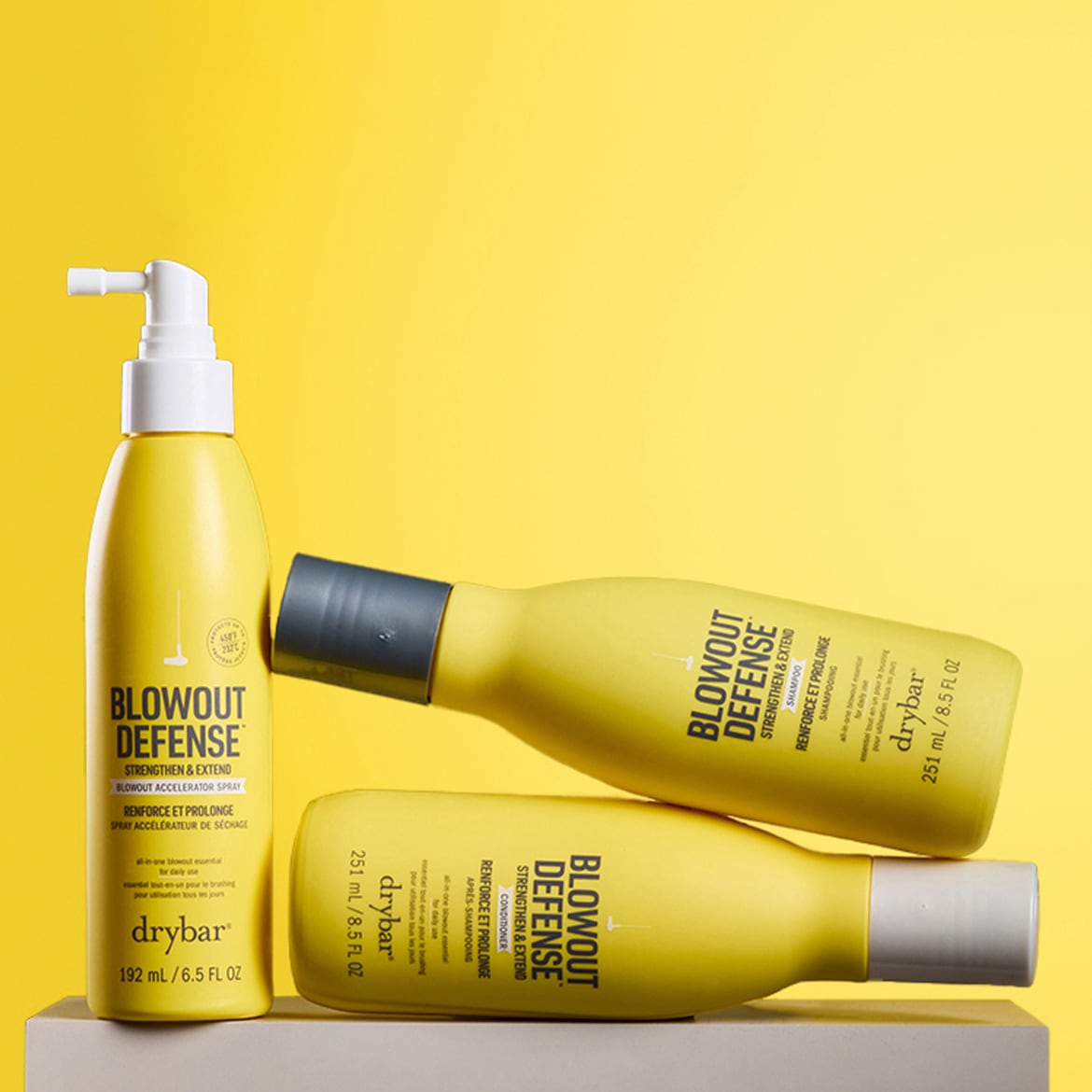 Drei gelbe Drybar Blowout Defense Produkte, darunter ein Blowout Accelerator Spray mit weißem Pumpspender, ein Shampoo und ein Conditioner, liegen auf einem hellen Untergrund vor einem leuchtend gelben Hintergrund.