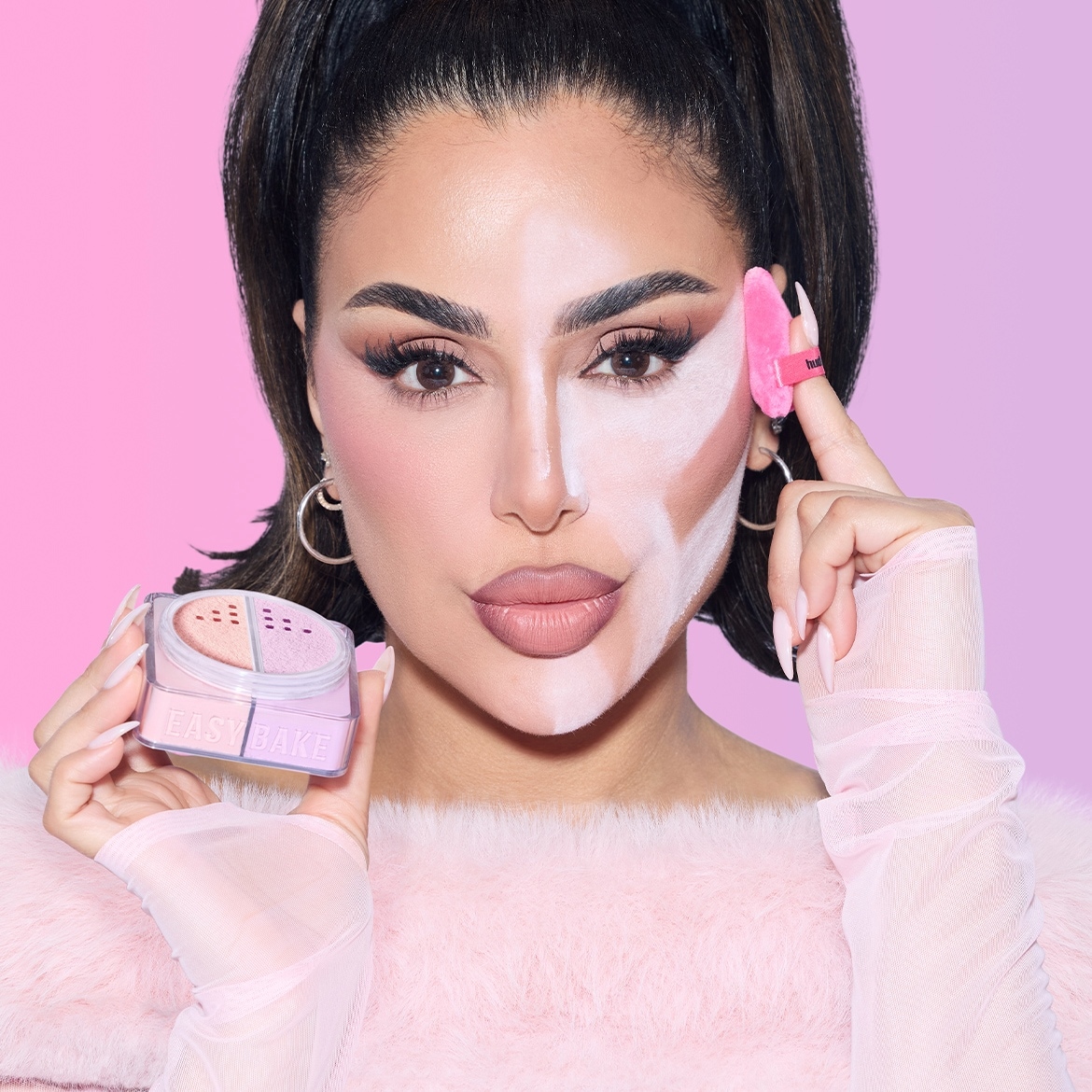 Eine Frau mit dunklen Haaren und Make-up auf der rechten Gesichtshälfte hält ein pinkes Puder und einen pinken Make-up-Schwamm in den Händen, während die linke Gesichtshälfte mit weißem Puder bedeckt ist.