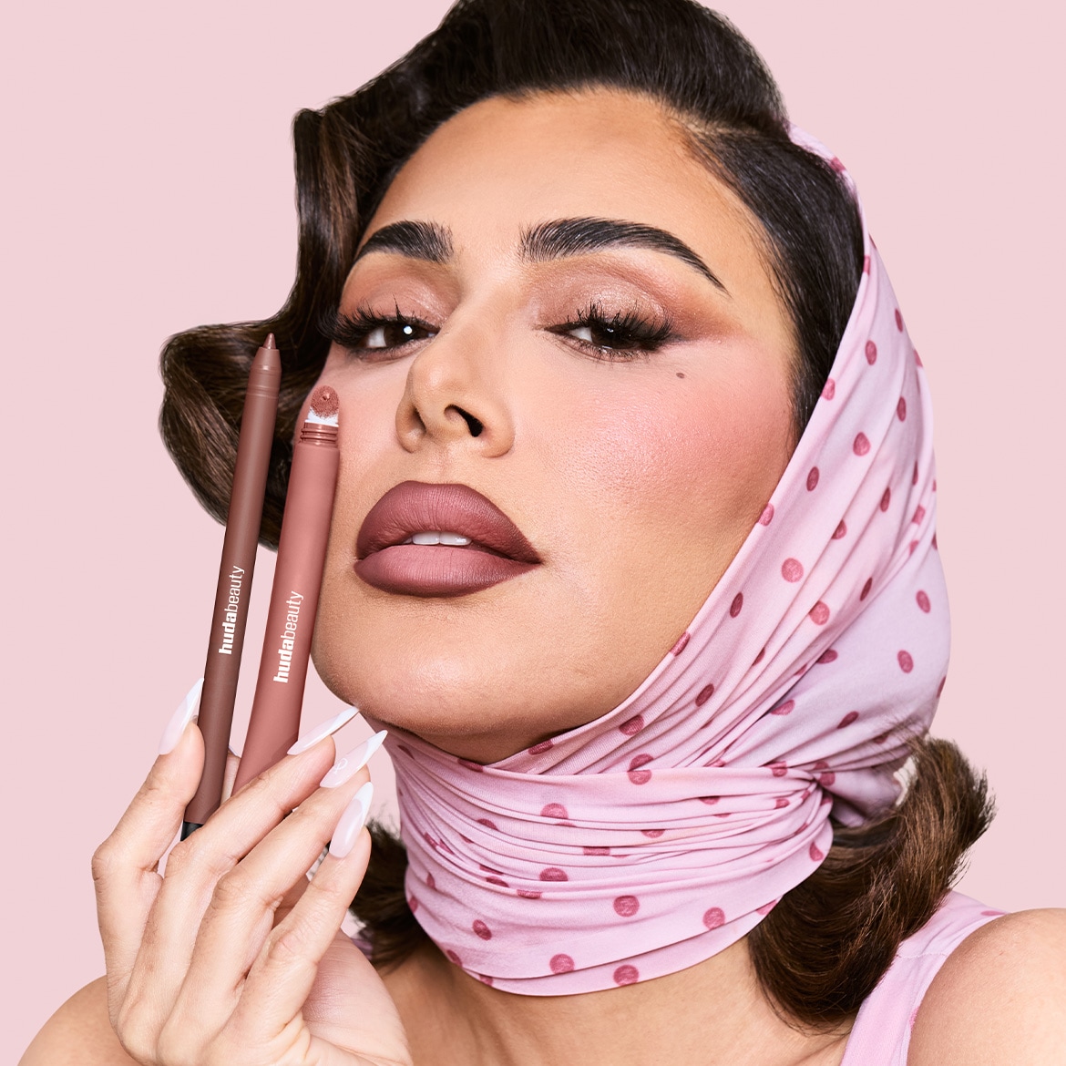 Eine Frau mit einem rosa gepunkteten Kopftuch hält zwei Huda Beauty Lippenprodukte in den Händen, einen Lipliner und einen flüssigen Lippenstift, die ihre perfekt geschminkten Lippen in einem matten Mauve-Ton ergänzen.