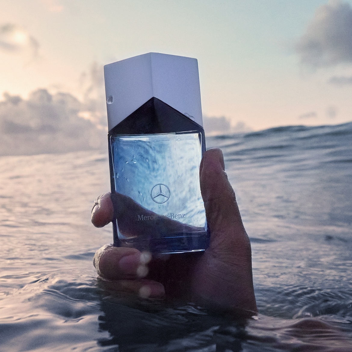 Eine Hand hält eine Flasche Mercedes-Benz-Parfüm im Wasser, wobei der Himmel und die Wellen im Hintergrund zu sehen sind.