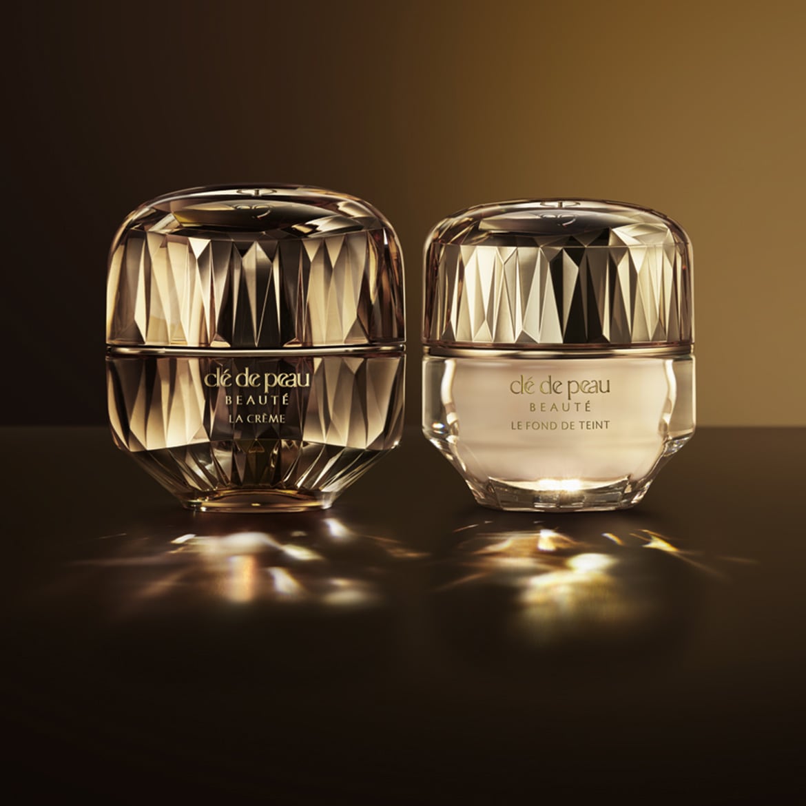 Zwei luxuriöse Tiegel von Clé de Peau Beauté, einer mit La Crème und der andere mit Le Fond de Teint, beide in eleganten, facettierten Goldverpackungen.