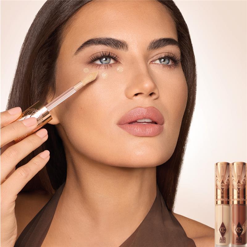 Nahaufnahme einer Frau, die mit einem Pinsel Concealer unter ihrem Auge aufträgt, mit zwei Charlotte Tilbury Airbrush Concealern im Hintergrund.