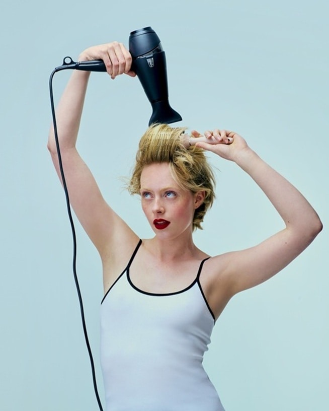 Eine Frau mit roten Lippen und weißem Top föhnt und bürstet ihre blonden Haare mit einem schwarzen Fön für ein perfektes Styling ohne Hitzeschäden.