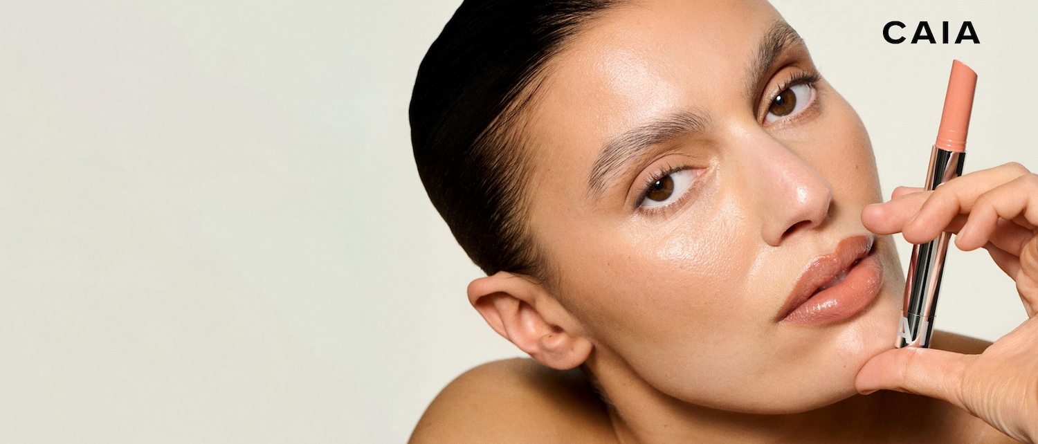 Nahaufnahme eines Models mit glänzenden Lippen, das einen CAIA-Lippenstift in der Hand hält, um einen strahlenden und natürlichen Look zu erzielen.
