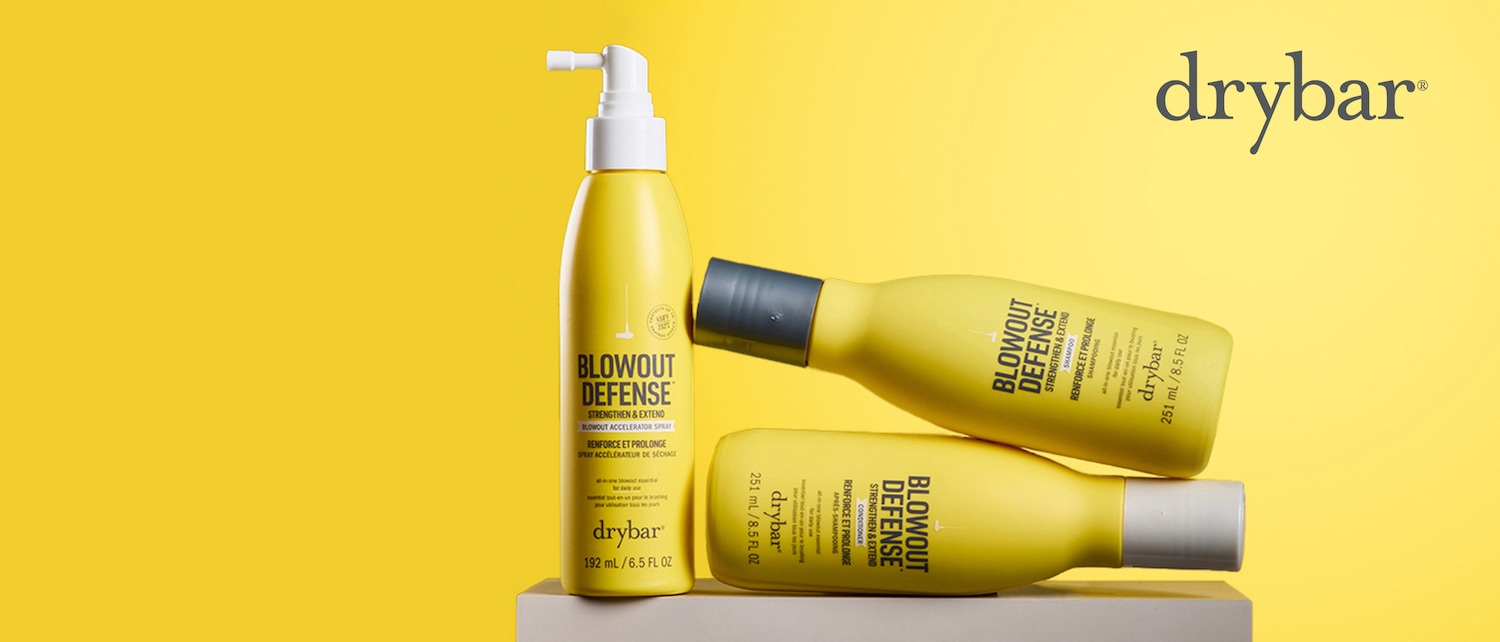 Drei gelbe Drybar Blowout Defense Flaschen, zwei liegen und eine steht aufrecht, präsentieren die "Strengthen & Extend" Serie, die Shampoo, Conditioner und Blowout Accelerator Spray umfasst.