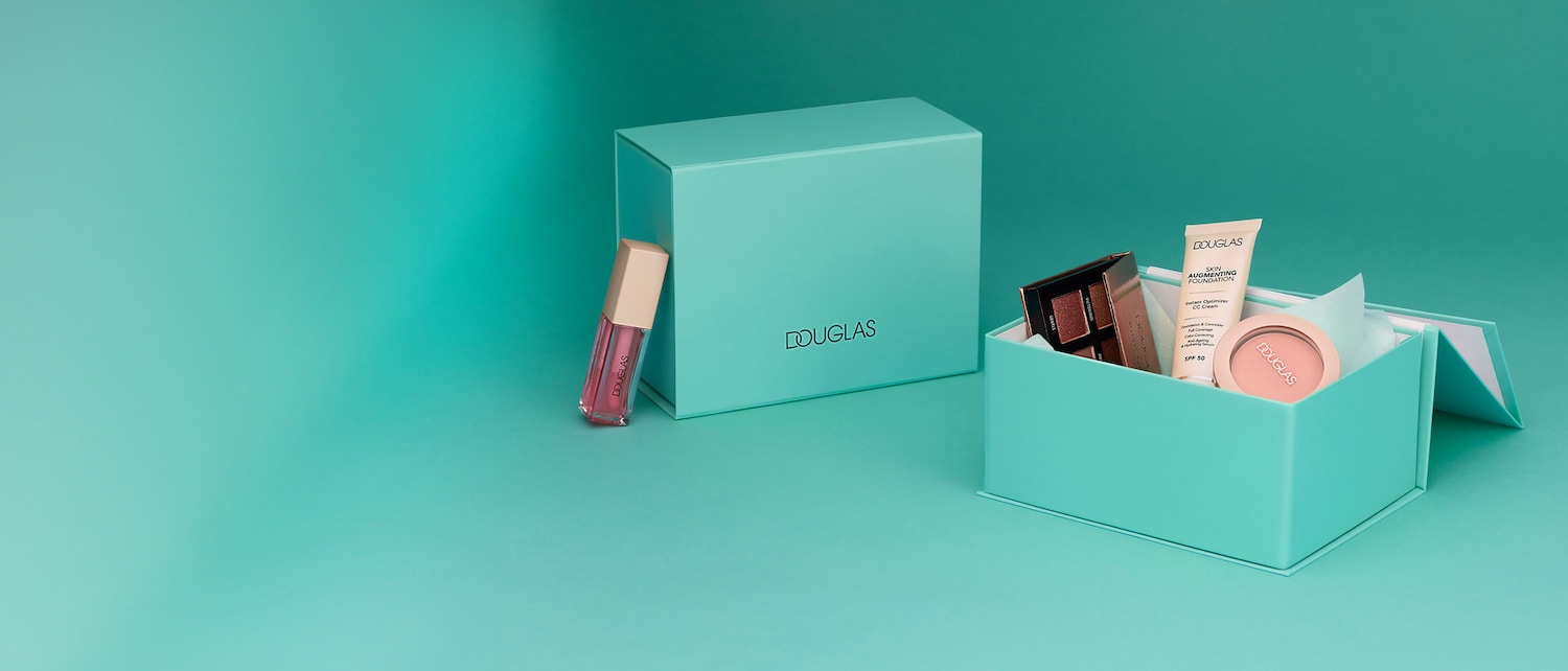 Eine Nahaufnahme zeigt eine Douglas-Geschenkbox in Türkis, die ein Douglas Skin Augmenting Foundation, eine Lidschattenpalette und einen Rouge enthält, daneben steht ein Douglas Lipgloss.