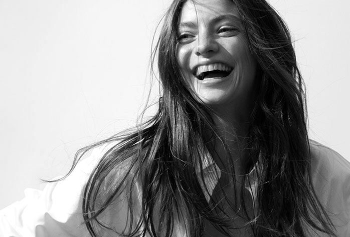 Schwarz-Weiß-Foto einer lachenden Frau mit langen, dunklen Haaren und einem hellen Hemd vor einem hellen Hintergrund.