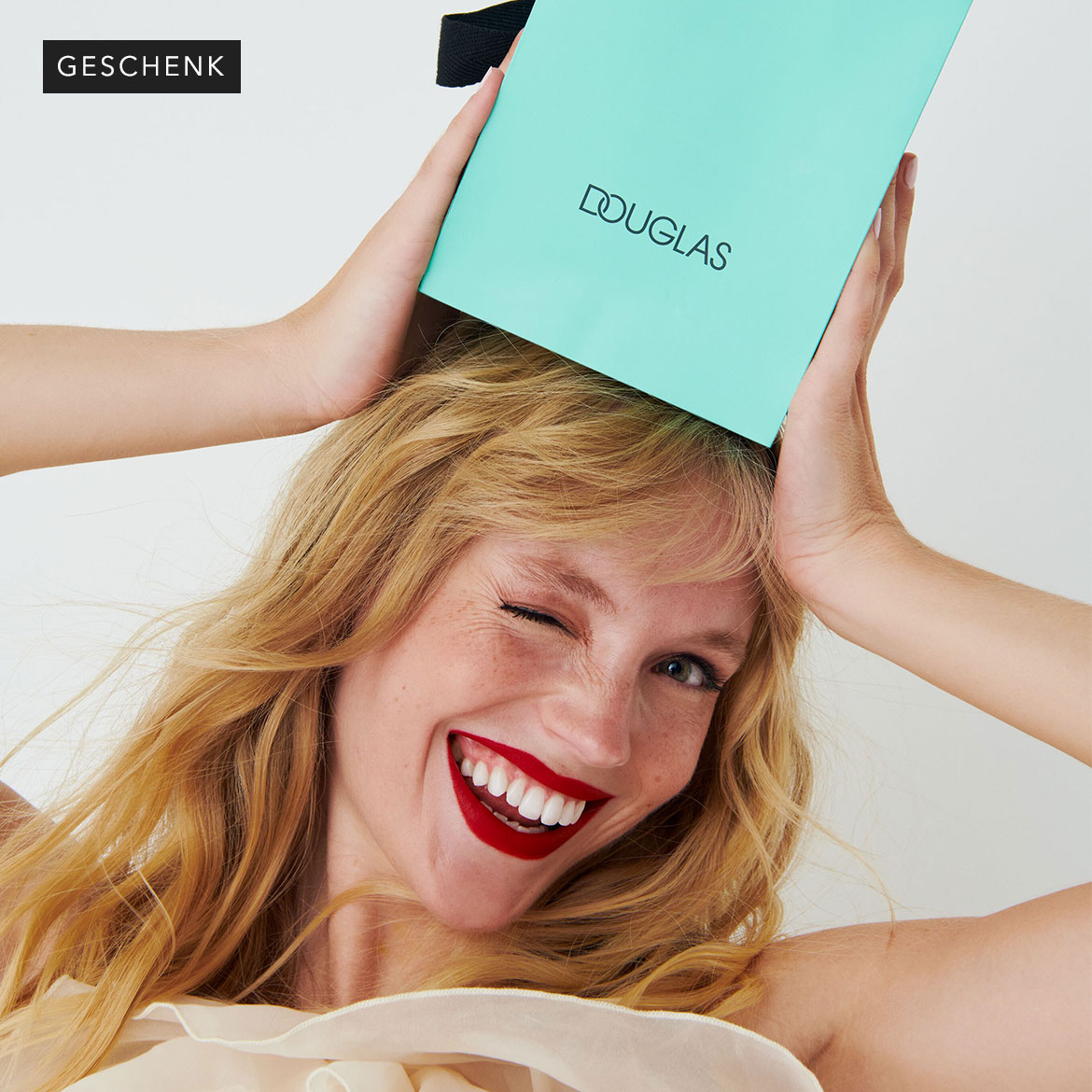Eine blonde Frau mit roten Lippen hält eine hellblaue Douglas-Geschenktüte über ihren Kopf und zwinkert.