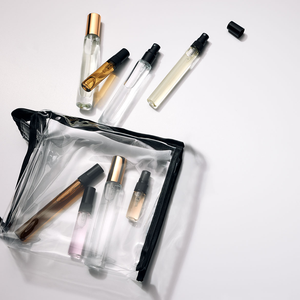 Eine durchsichtige Kosmetiktasche mit Reißverschluss enthält mehrere Parfümproben mit goldenen, schwarzen und silbernen Akzenten, einige davon liegen daneben.