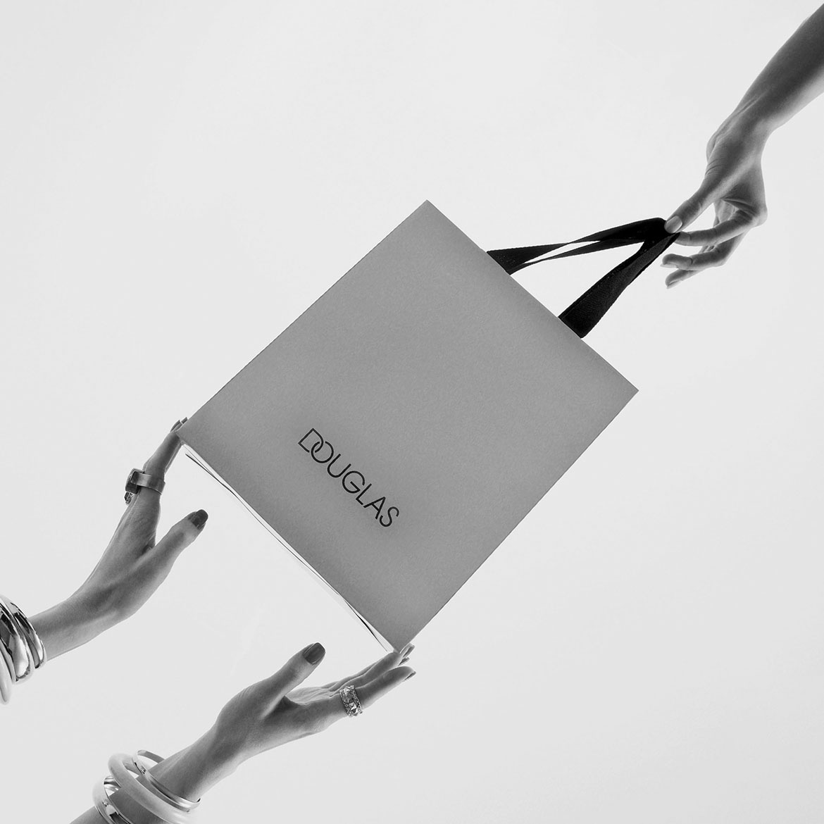 Eine Douglas-Geschenktüte in Grau wird von zwei Händen mit Schmuck gehalten, die zum Einkaufen einladen.