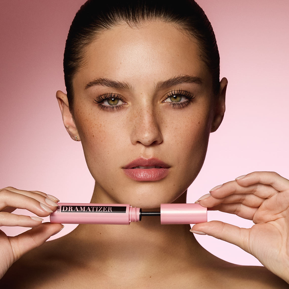 Eine Frau mit grünen Augen und Sommersprossen hält eine offene pinkfarbene Mascara mit der Aufschrift „DRAMATIZER“ in ihren Händen, wobei die Bürste sichtbar ist.
