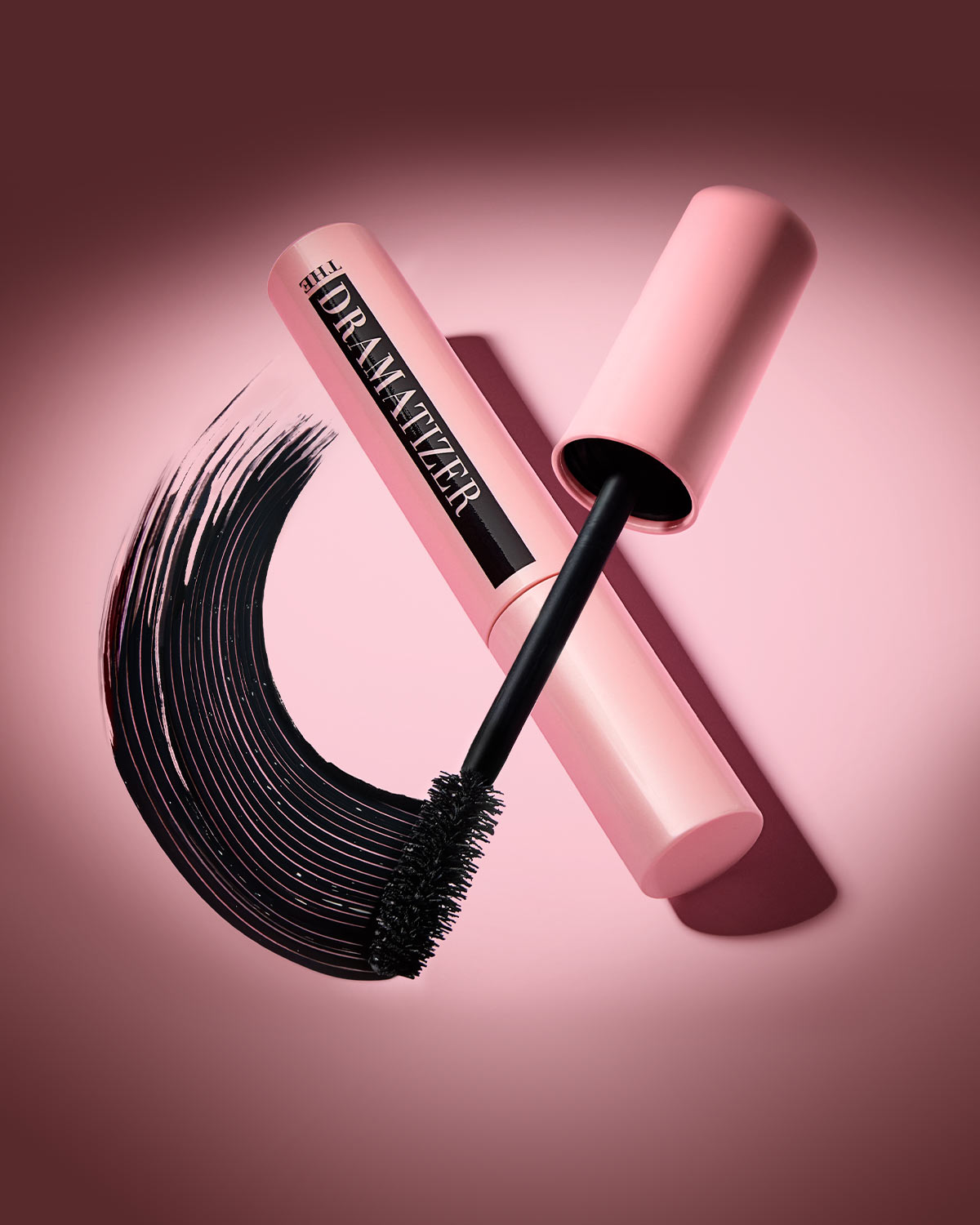 Die Dramatizer Mascara in einem hellrosa Behälter mit schwarzem Schriftzug „THE DRAMATIZER“ und einer Bürste, die einen geschwungenen schwarzen Mascara-Strich auf einem rosa Hintergrund hinterlässt.
