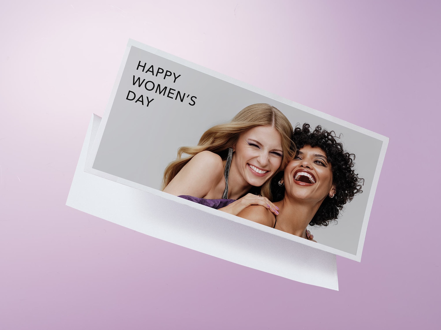 Eine Karte mit der Aufschrift "Happy Women's Day" zeigt zwei lachende Frauen, eine mit blonden, die andere mit lockigen dunklen Haaren, vor einem zartrosa Hintergrund.
