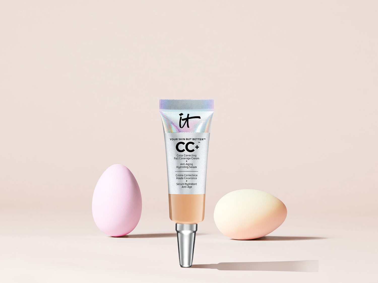 Close-up of IT Cosmetics CC+ cream tube with a holographic cap, surrounded by pink and peach-colored blending sponges, promising color correction, full coverage, anti-aging, and hydration.
