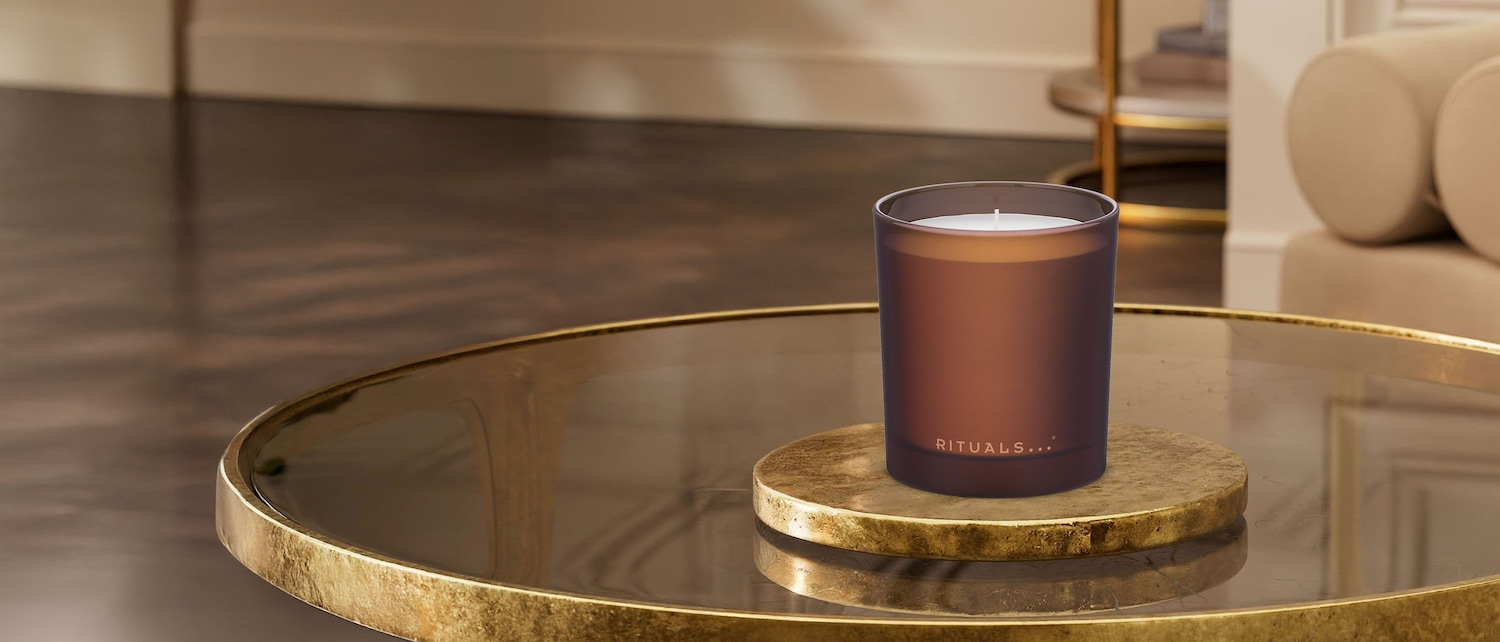 Ein braunes Rituals... Duftkerzenglas steht auf einem goldenen Untersetzer auf einem goldenen Tisch.