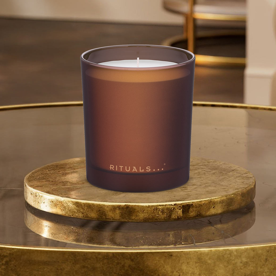Ein braunes Rituals... Duftkerzenglas steht auf einem goldenen Untersetzer und verströmt einen angenehmen Duft.