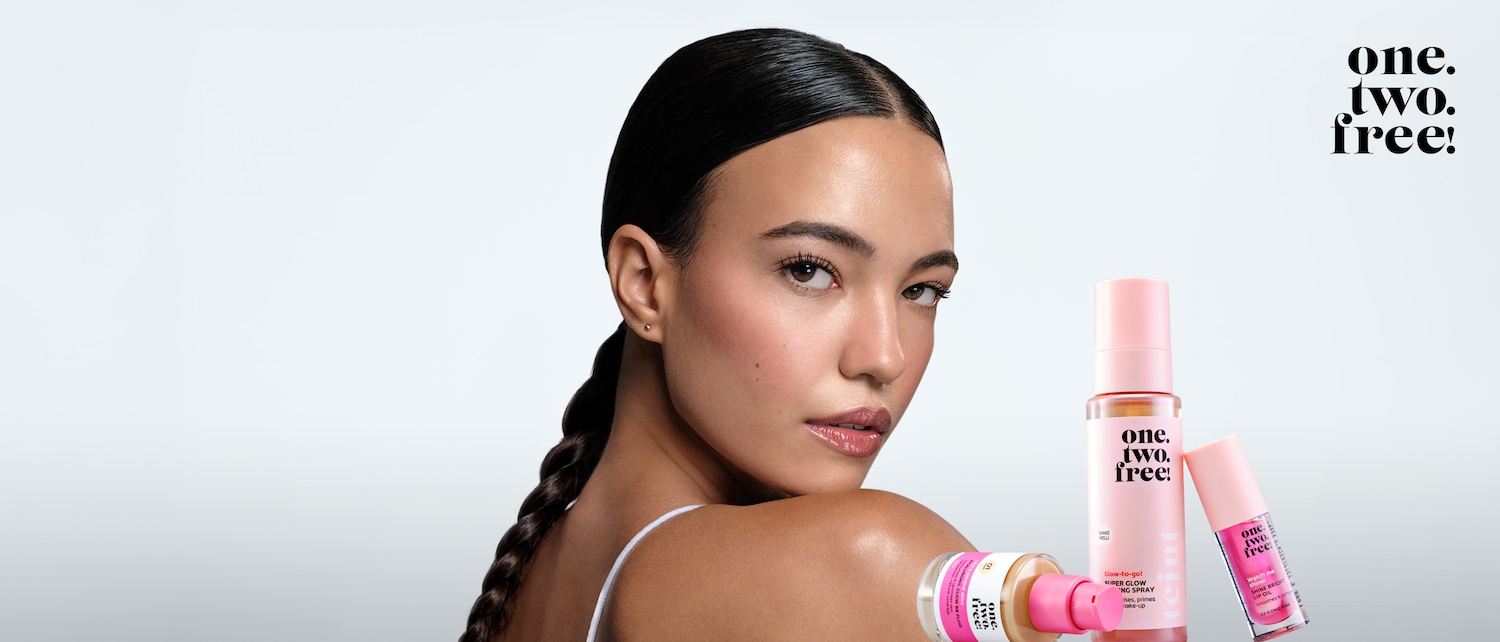 Eine Frau mit glänzender Haut und zurückgekämmtem Haar posiert neben Produkten von One.Two.Free!, darunter ein Glow BB Fluid, ein Super Glow Fixing Spray und ein Shine Bright Lip Oil.