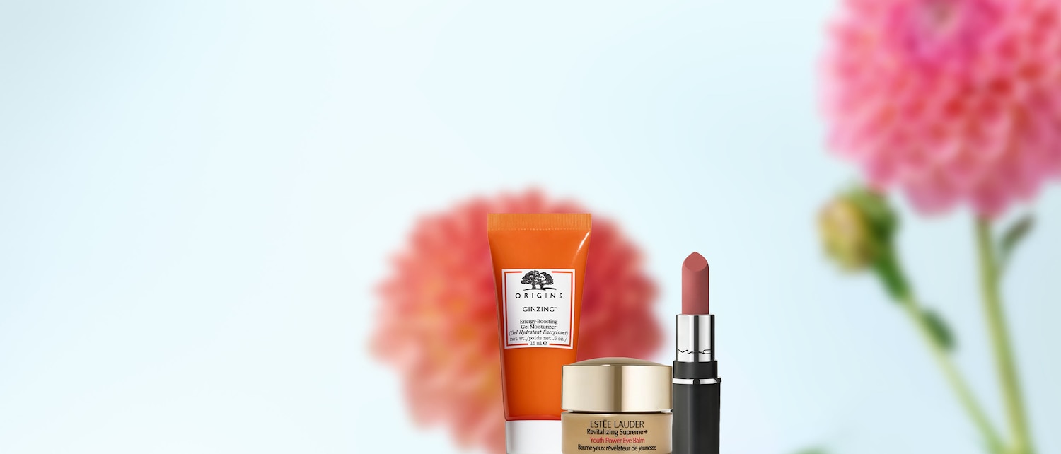 Vor einem hellblauen Hintergrund sind Schönheitsprodukte wie Origins Ginzing Feuchtigkeitsgel in einer orangefarbenen Tube, Estée Lauder Augenbalsam in einem goldenen Tiegel und ein MAC Lippenstift in einem silbernen Gehäuse zu sehen.