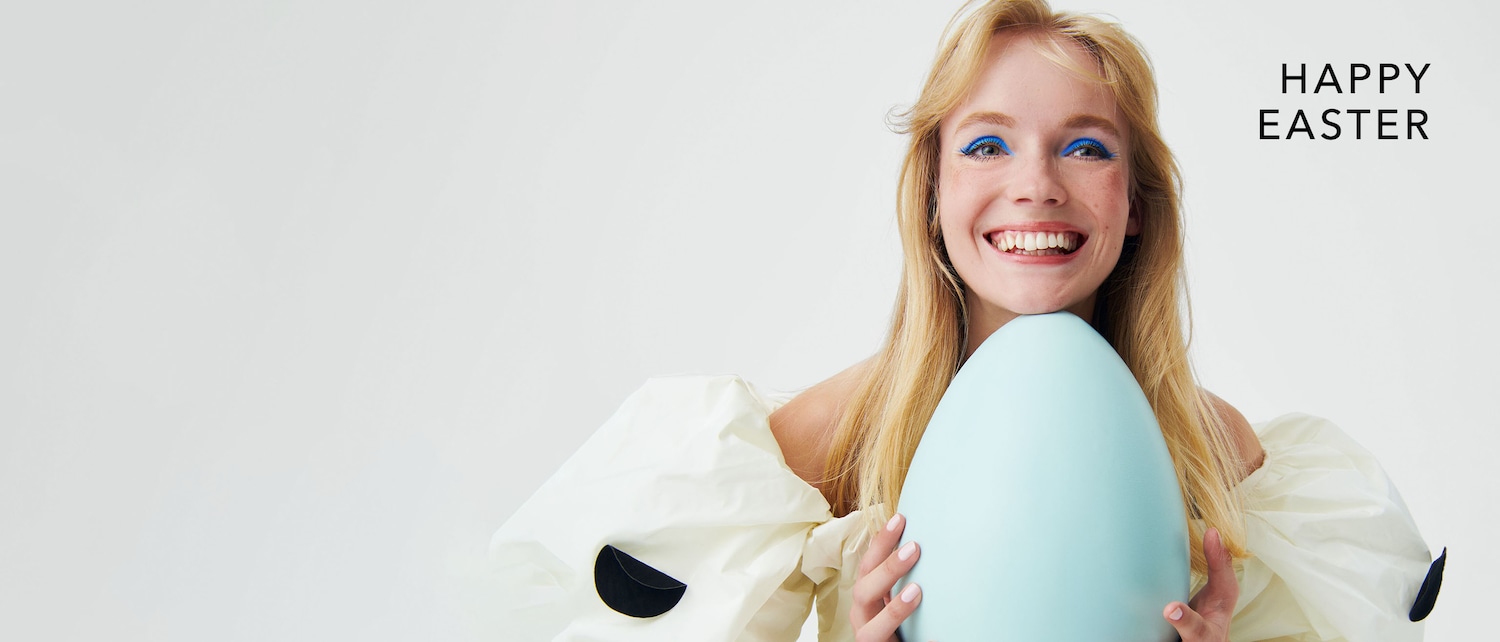 Fröhliche Ostern! Eine strahlende Frau mit blonden Haaren und blauem Eyeliner umarmt ein hellblaues Osterei und wünscht Ihnen ein frohes Fest.