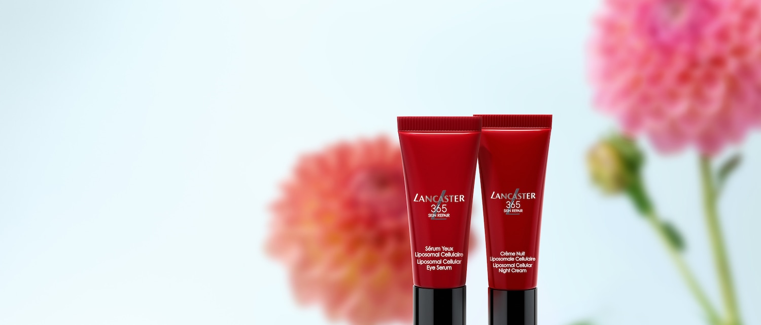 Zwei rote Tuben Lancaster 365 Skin Repair, eine mit Augenserum und die andere mit Nachtcreme, stehen vor einem verschwommenen rosa Blumenhintergrund.
